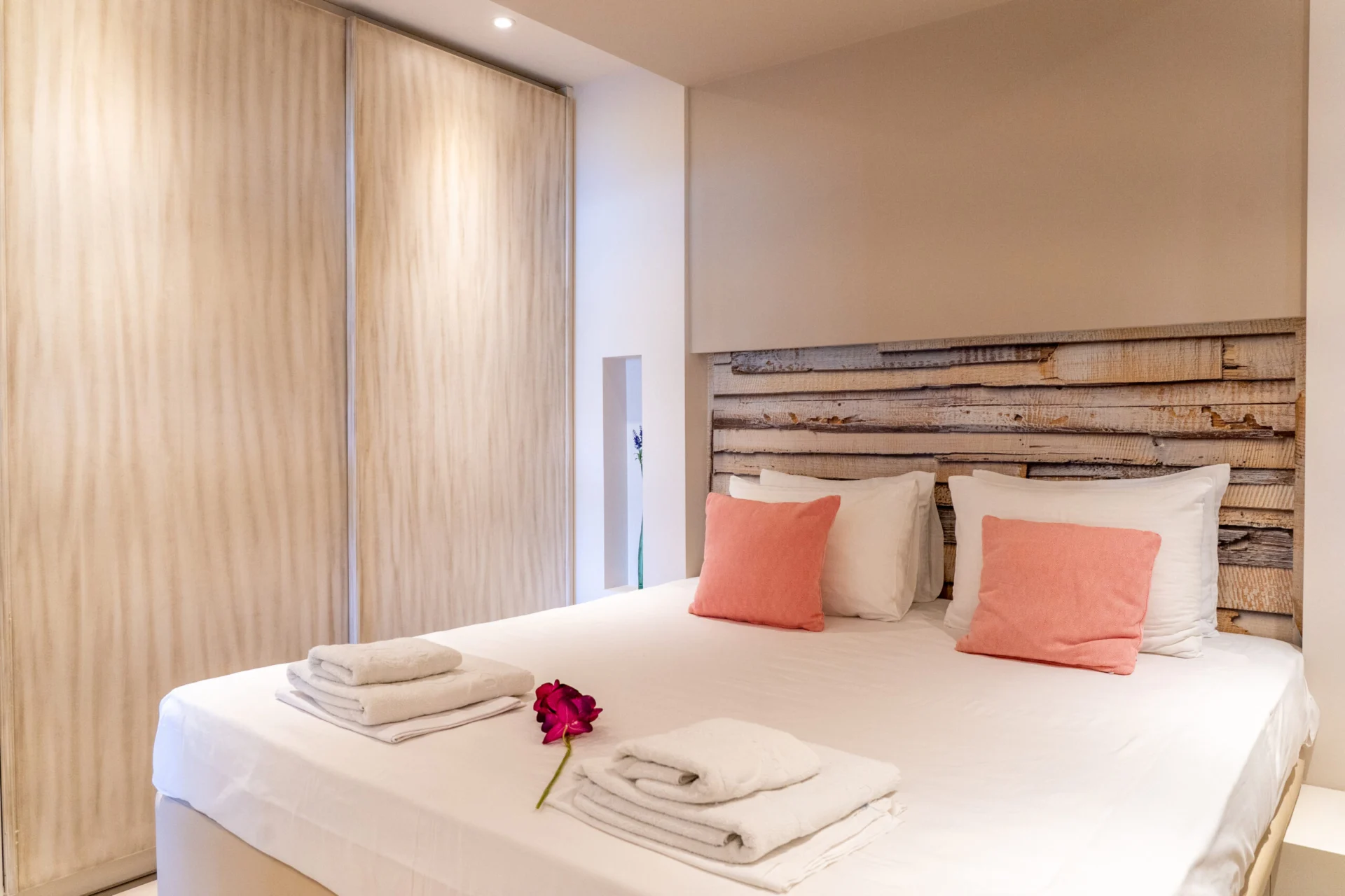 A bed with a reclaimed-wood headboard, coral cushions and a sliding wardrobe in the Kassandra Signature Suite at CORE Luxury Suites, Skiathos.