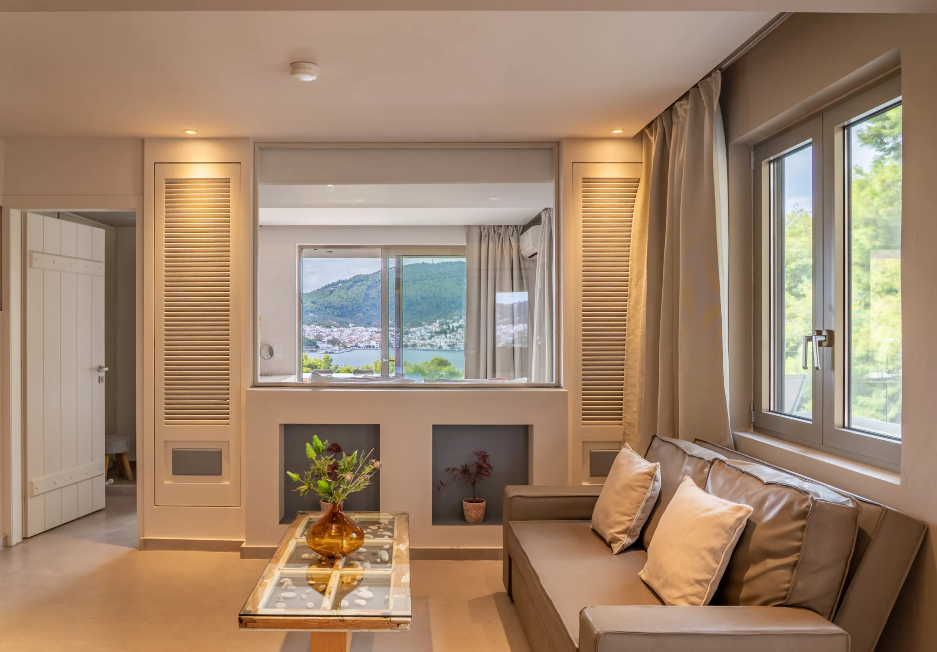 A living room with a sofa, a glass table and the view through the window in the Stavros Family Suite at CORE Luxury Suites, Skiathos.