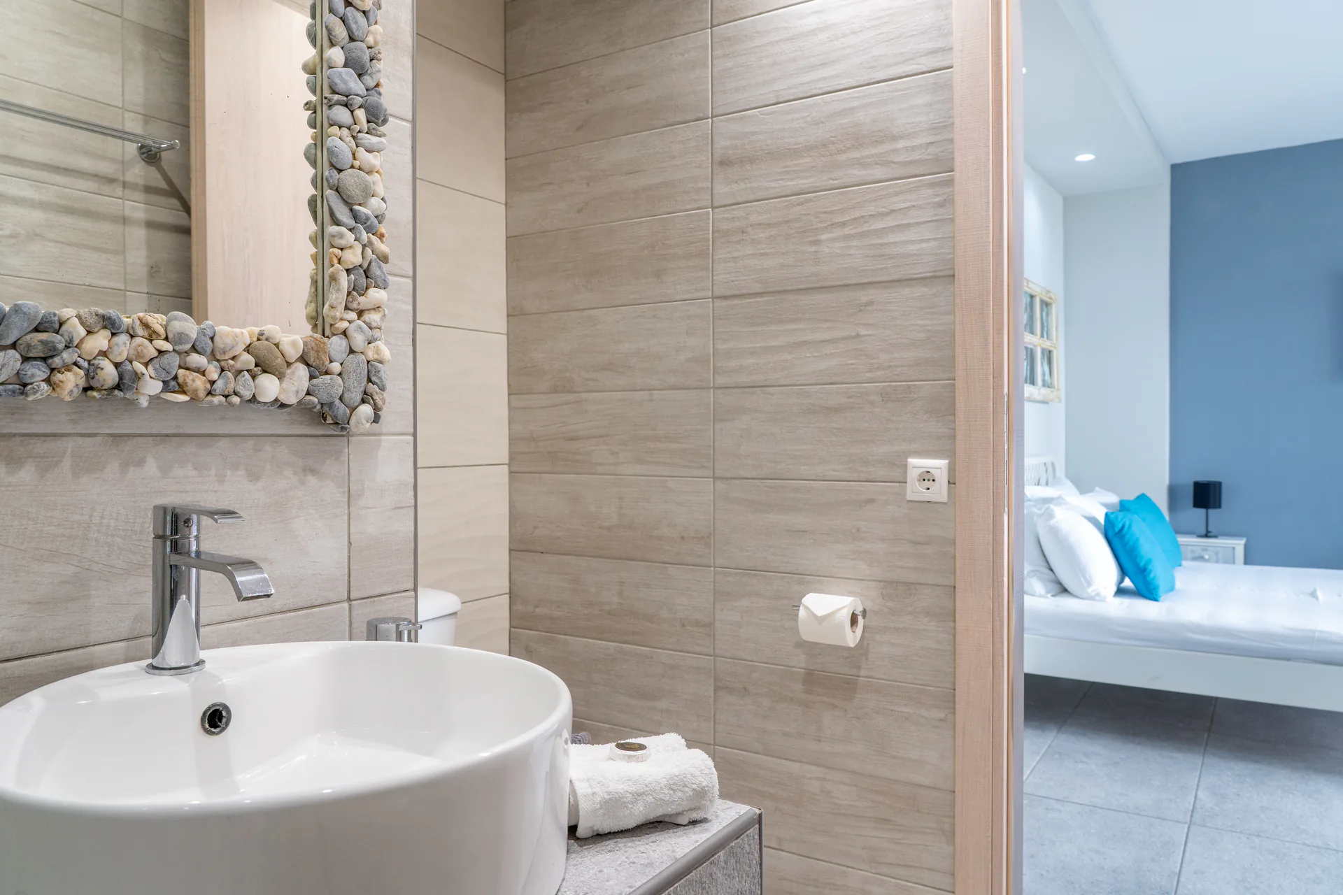 Un lavabo e uno specchio incorniciato da ciottoli, con la camera visibile attraverso la porta, nella Alex 1 Classic Suite di CORE Luxury Suites, Skiathos.