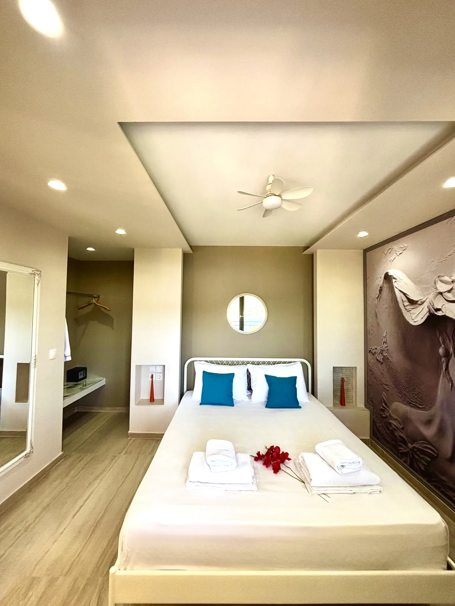 Ein Bett mit petrolfarbenen Kissen unter einem runden Spiegel und geschnitzten Paneel in der Cassy Deluxe Suite bei CORE Luxury Suites, Skiathos.