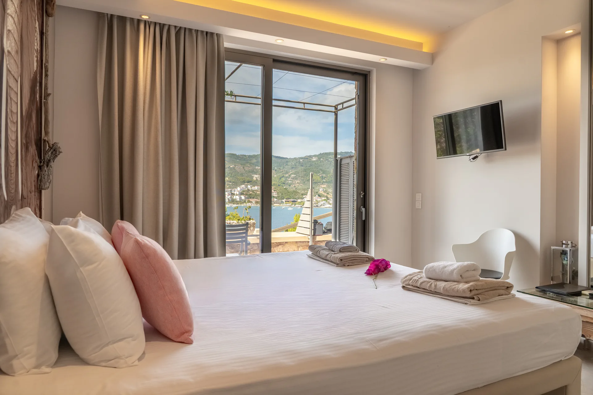 A bed with pink cushions facing doors open to the bay in the Maggi Superior Suite at CORE Luxury Suites, Skiathos.